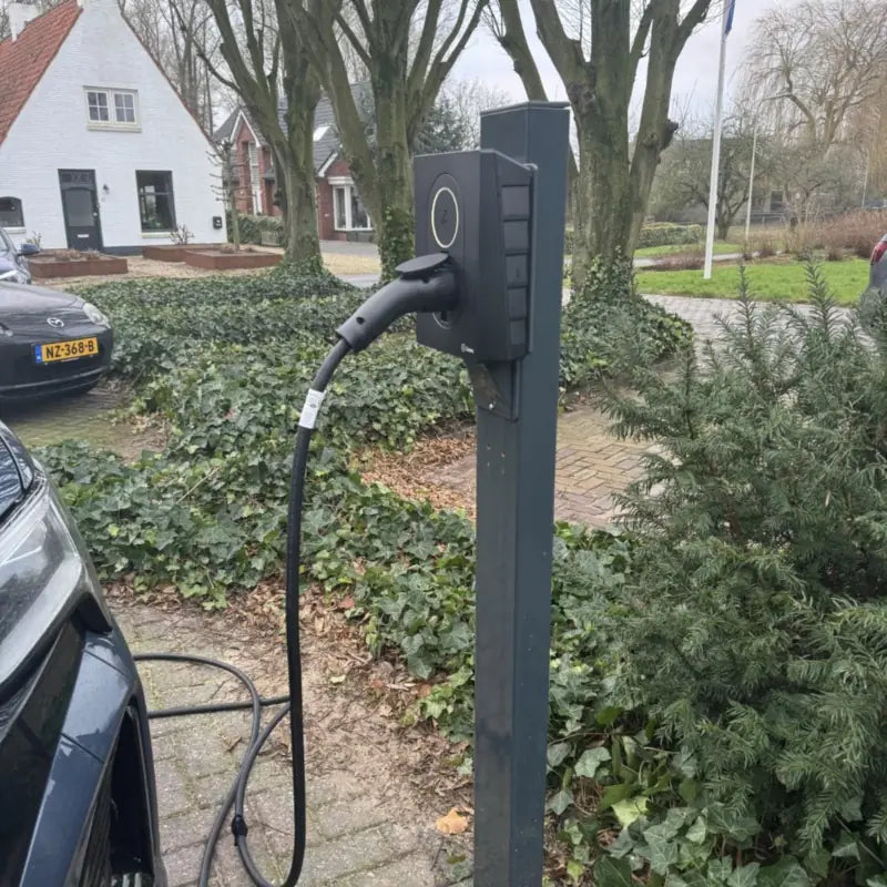 Zaptec Go laadpaal op een donkergrijze paal bij een oprit, met een aangesloten laadkabel naar een elektrische auto.
