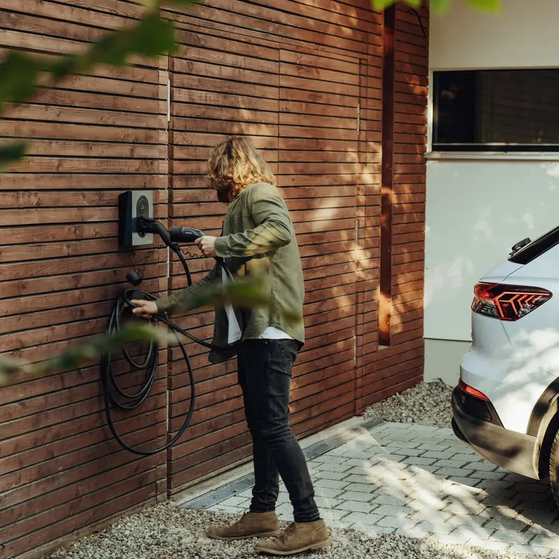 Man pakt de Type 2-laadkabel van een Zaptec Go laadpaal aan een houten gevel om zijn elektrische SUV op te laden.