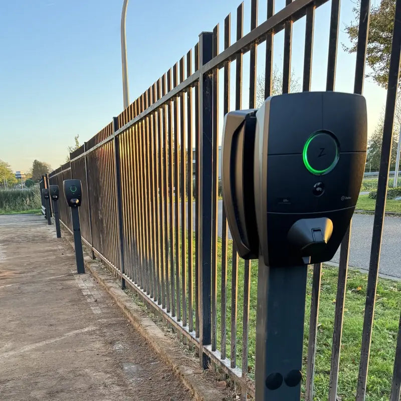 Rij Zaptec Pro laadpalen op palen langs een stalen hekwerk op een bedrijfsparkeerplaats, met groene statusringen.